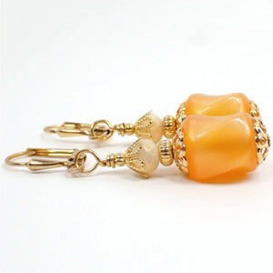 Side view of the handmade drop earrings. The metal is gold plated in color. There are new faceted glass crystal beads at the top that are off white with a light golden sheen on them. The bottom beads are vintage moonglow lucite beads in peach color. They are twisted barrel shaped and have an inner glowy like appearance as you move around in the light.