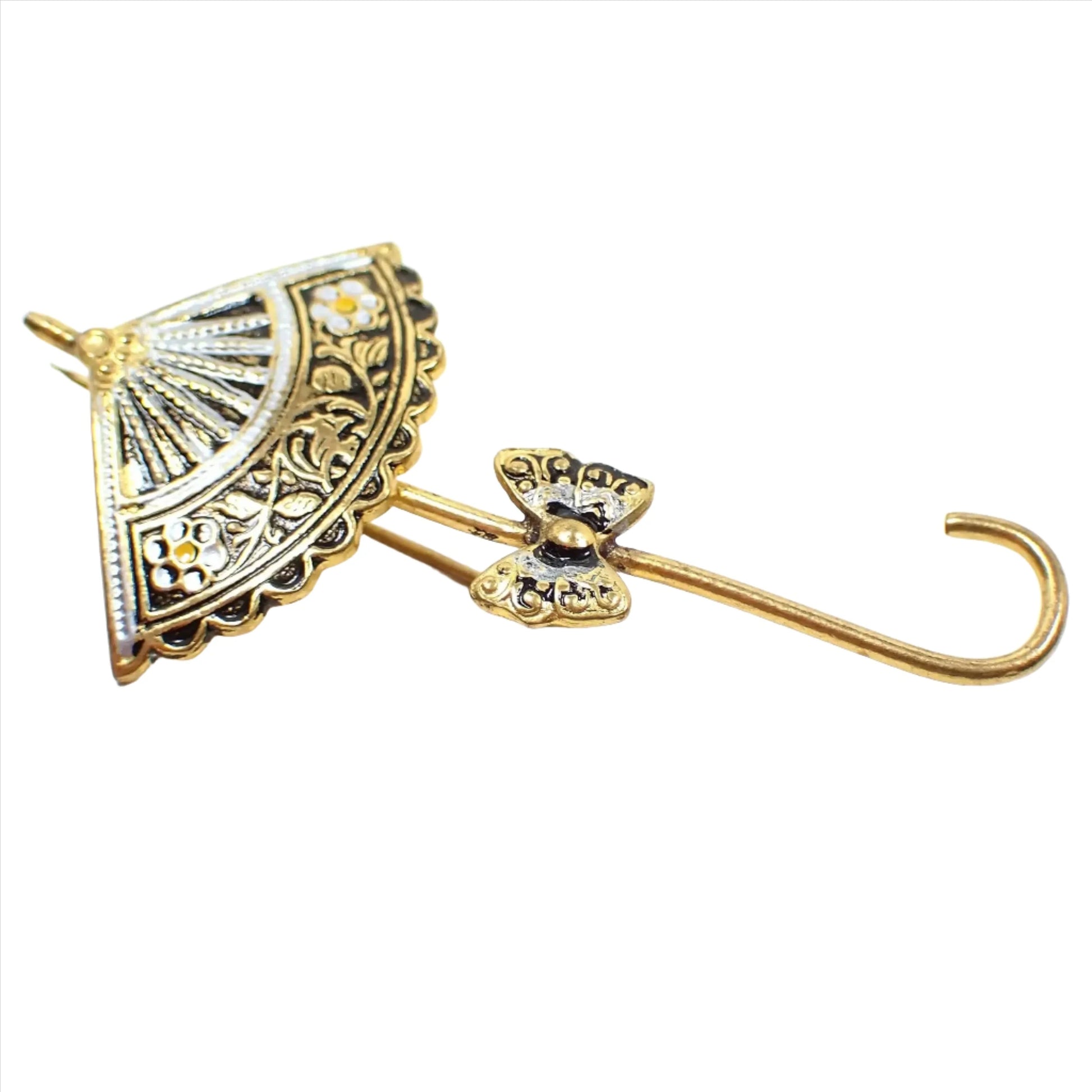 Enlarged view of the Mid Century vintage Damascene brooch pin. It is shaped like an umbrella with a butterfly on the upper handle area. The metal is gold tone plated in color and it has a gold tone, metallic silver painted, and black painted Damascene style design.