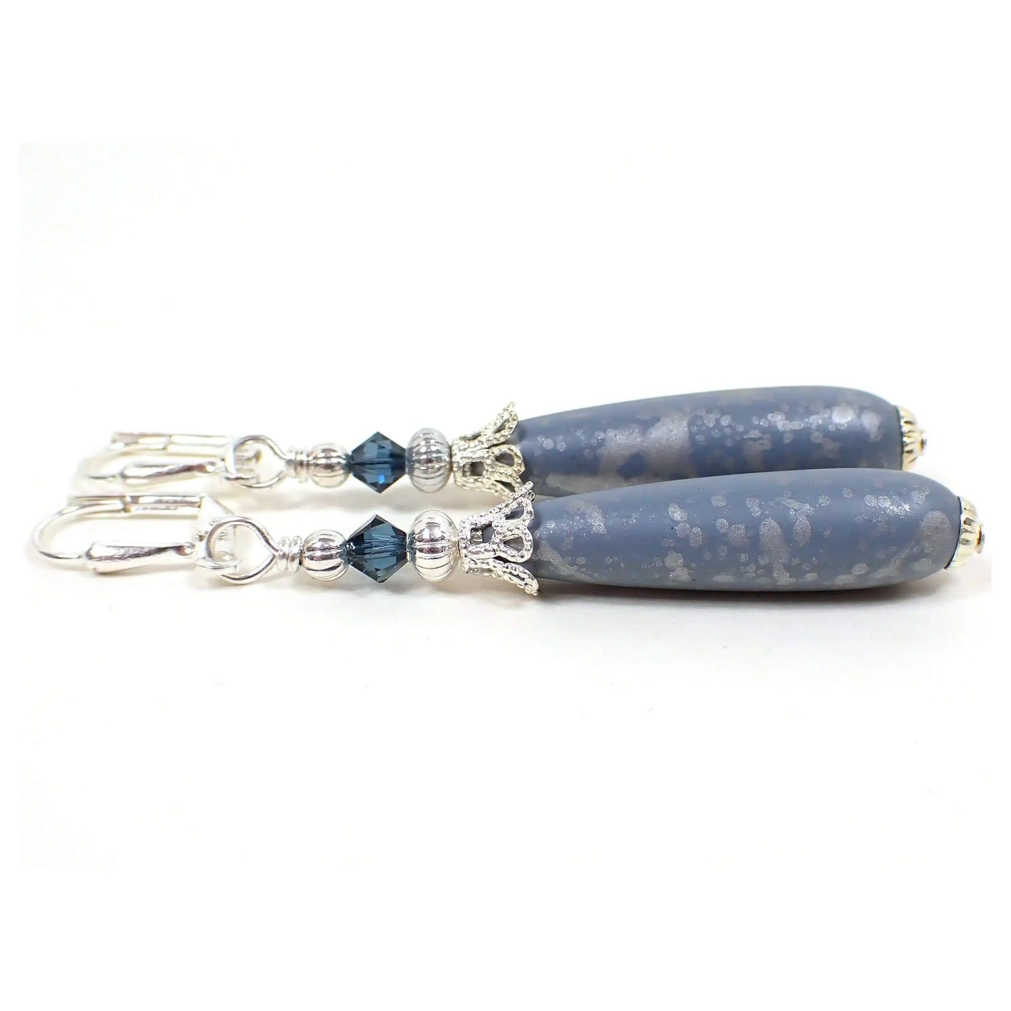 Side view of the handmade teardrop earrings. The metal is silver plated in color. There are blue faceted glass crystal beads on the top. The bottom beads are acrylic and teardrop shaped. They are a light country blue color with a metallic silver splash pattern on them.