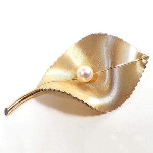 Front view of the Mid Century vintage Krementz brooch pin. The metal is a matte brushed textured gold tone plated in color. The brooch is shaped like a curvy leaf with a stem and scalloped edges on the sides. There is a cultured pearl in the middle of the leaf.