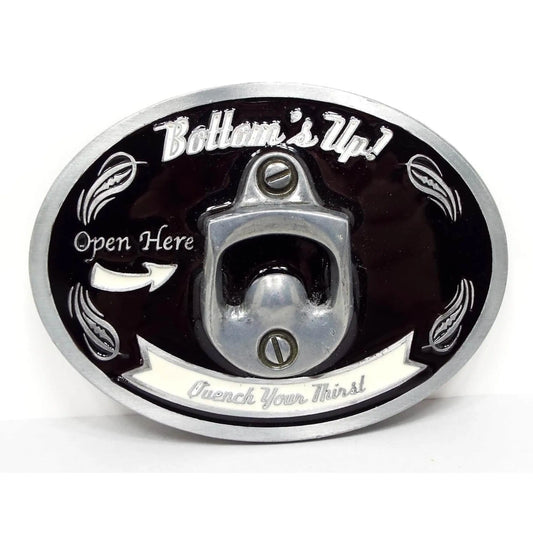 Front view of the retro vintage Bottom's Up belt buckle. It is oval and silver tone in color with black enamel and white enameled design and lettering. It says Bottom's Up, Open Here, and Quench your Thirst. There is a bottle opener in the middle of the buckle.