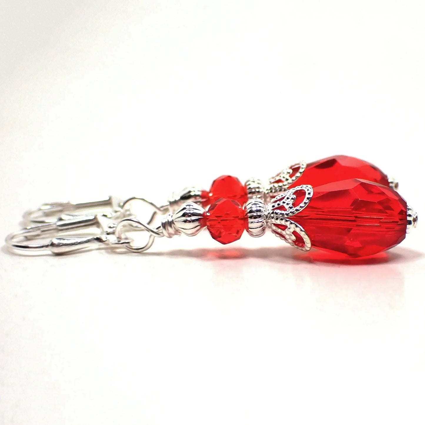 Angled view of the Victorian style handmade crystal glass teardrop earrings. The metal is silver plated in color. There is a faceted rondelle bead at the top and a faceted teardrop bead at the bottom. Both beads are a bright red color.