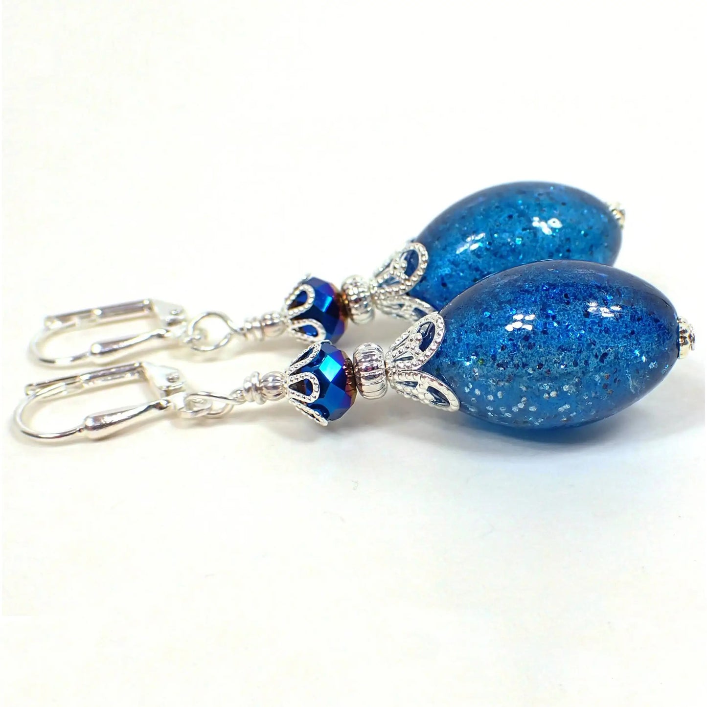 Angled view of the handmade drop earrings. The metal is silver plated in color. There are metallic blue faceted glass beads at the top. The bottom beads are acrylic ovals in a semi translucent bright blue color. They have blue glitter embedded in them.