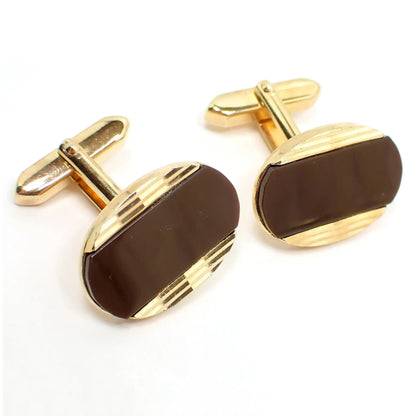 Angled front view of the retro vintage lucite cufflinks. The metal is gold tone plated in color. They are oval shaped with cut engraved lines at the top and bottom. The middle area has pearly brown lucite cab insets.