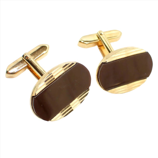 Angled front view of the retro vintage lucite cufflinks. The metal is gold tone plated in color. They are oval shaped with cut engraved lines at the top and bottom. The middle area has pearly brown lucite cab insets.