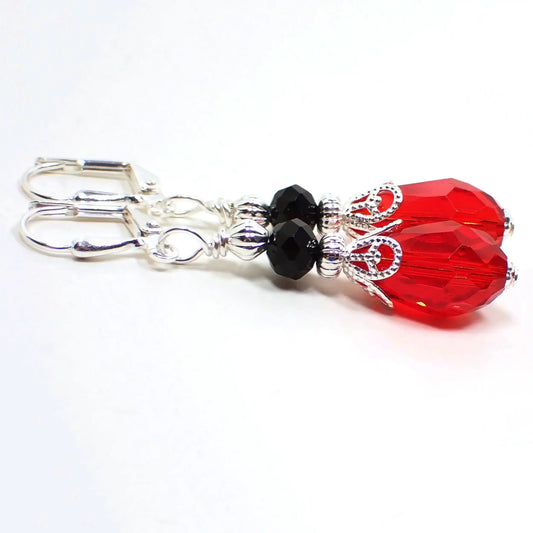 Angled view of the Victorian style handmade crystal glass teardrop earrings. The metal is silver plated in color. There is a faceted rondelle bead at the top in black and a faceted teardrop bead in red the bottom.