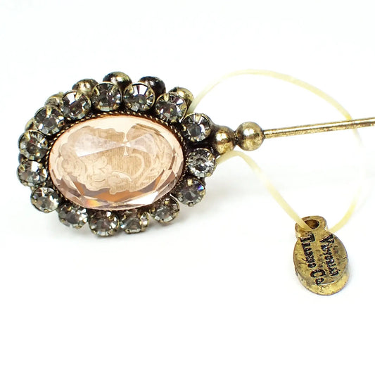 Enlarged angled top view of the pre owned Victorian Trading Company hat pin. The metal is antiqued brass in color. There is an oval faceted glass crystal cameo at the top in light pink that has a ladies head design. It is surrounded by round clear rhinestones on the edge. There is a hang tag attached with rubber string that says Victorian Trading Co on it.