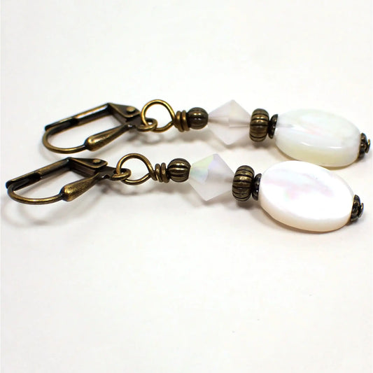 Angled view of the handmade mother of pearl drop earrings. The metal is antiqued brass in color. There are faceted AB frosted glass crystal beads at the top. The bottom mother of pearl shell beads are flat oval and have pearly areas that shimmer as you move around in the light.