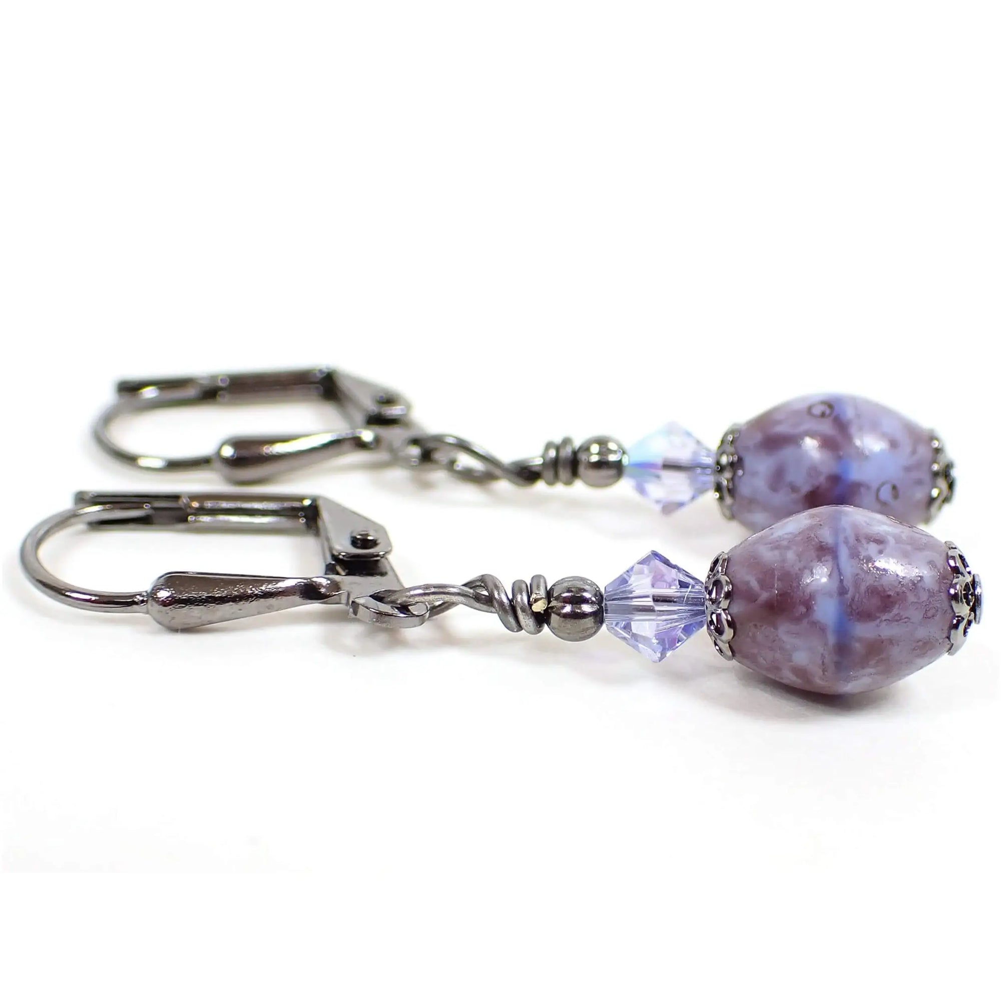 Side view of the handmade drop earrings with Czech beads. The metal is gunmetal gray in color. There are small faceted glass crystal beads at the top in a light purple color. The bottom Czech beads are glass, oval shaped, and have a mottled design with shades of purple on them.