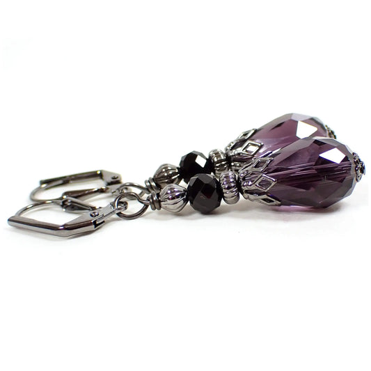 Side view of the handmade faceted glass crystal beaded earrings. The metal is dark gunmetal gray in color. There is a faceted black bead at the top and a faceted dark purple teardrop bead at the bottom.