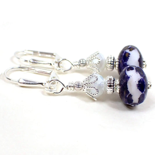 Side view of the handmade glass beaded drop earrings. The metal is silver plated in color. there are pearly opaque white faceted glass beads at the top. The bottom bead is a faceted rondelle shape and has dark blue and white pattern with tiny flecks of metallic antiqued gold paint.