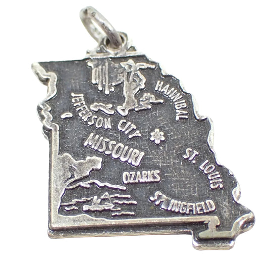 Enlarged view of the retro vintage sterling silver souvenir charm. It's shaped like the state of Missouri and has several cities on it, as well as depictions of notable things. The sterling has darkened for an antiqued silver color of silver and dark gray. There is a small jump ring attached to the top.