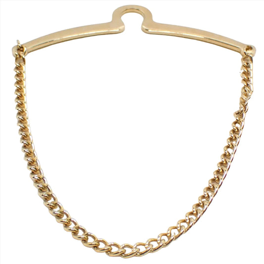 Enlarged front view of the retro vintage tie chain. The metal is gold tone plated in color. There is a curved bar at the top with a curve in the middle to go over a button. The chain is curb link and hangs down from one side to the other.