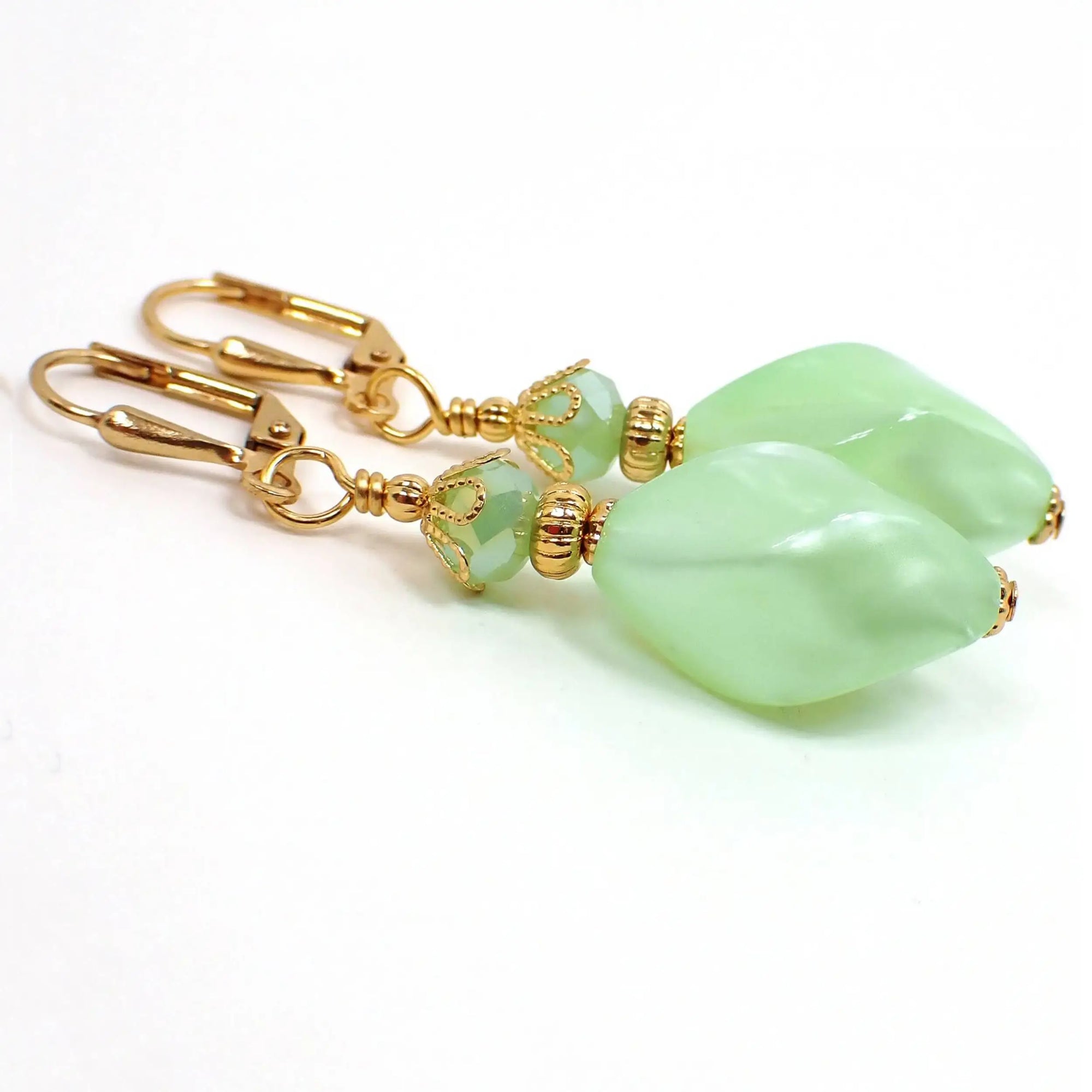 Angled view of the handmade twist drop earrings with vintage lucite beds. The metal is gold plated in color. There are new faceted light green glass beads at the top. The bottom lucite beads are pearly light green in color and have an angled twist like design.