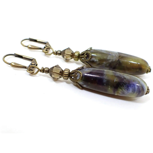 Side view of the handmade earrings with long vintage lucite beads. The metal is antiqued brass in color. There are faceted brown glass crystal beads at the top. The bottom lucite beads are long ovals and have swirled colors. Some of the colors seen are olive green, white, blue, gray, and brown. 