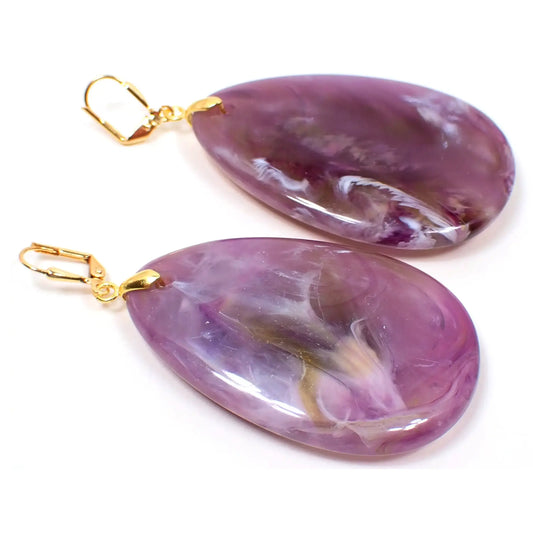 Angled view of the big heavy handmade teardrop earrings. The metal is gold plated in color. They are large teardrop shaped with shades of purple that has swirls of white and brown marbled in.