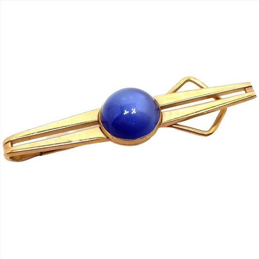 Angled front view of the Swank 1940s Mid Century vintage tie bar. The metal is gold tone in color and it has a Modernist design with open middle and tapered ends. There is a domed round moonglow lucite cab in the middle that is a rich blue in color.