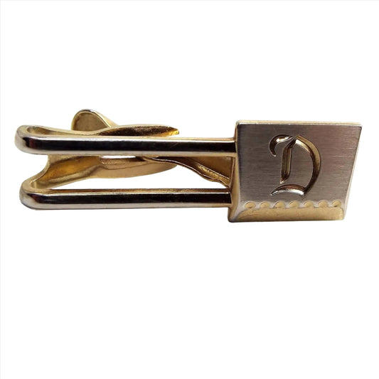 Front view of the Mid Century vintage Hickok initial tie clip. It has an open bar area with an angled square at the end. There is a letter D engraved on the front. The front part of the square is silver tone in color and the rest of the tie clip is gold tone in color.
