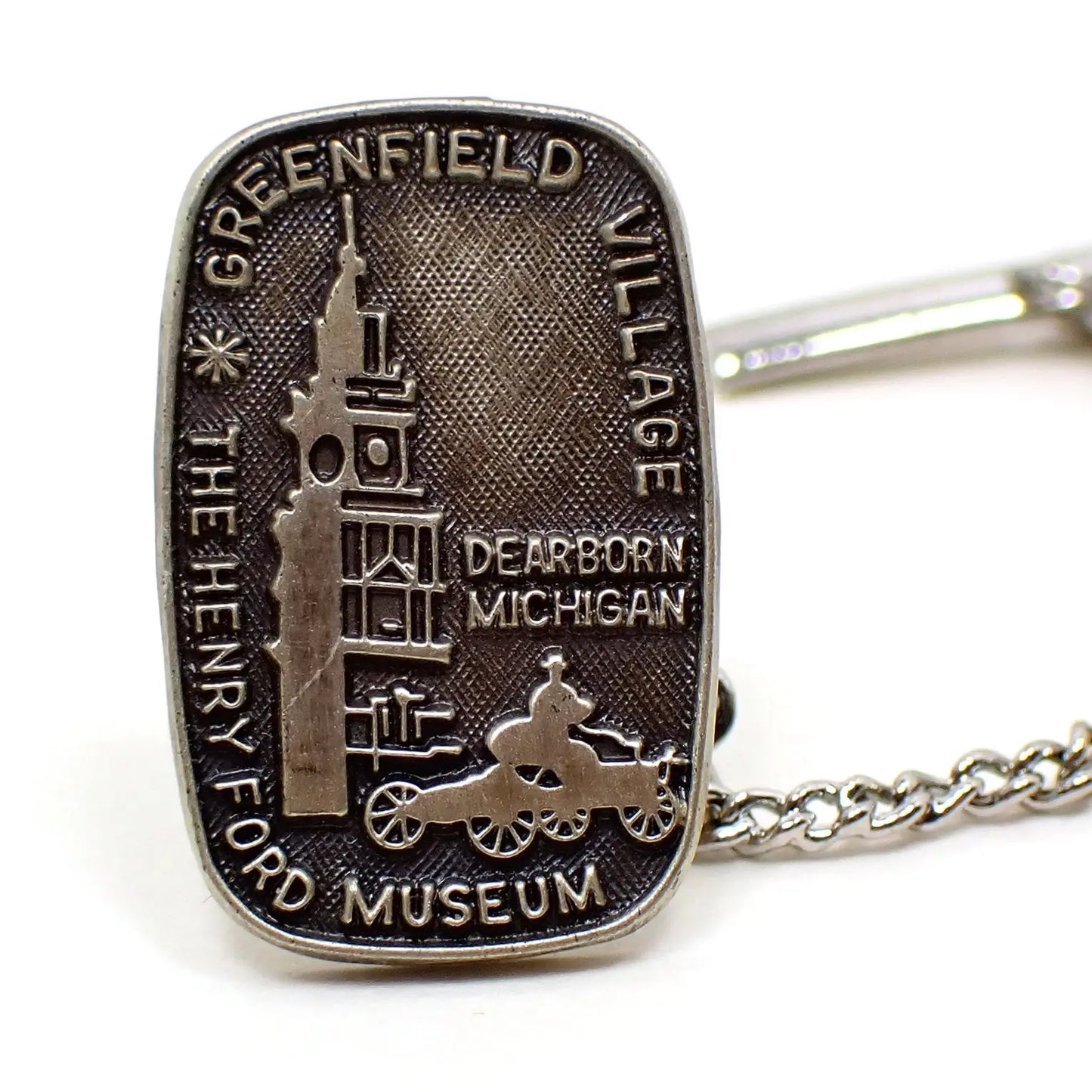 Enlarged front view of The Henry Ford Museum tie tack lapel pin. It has a rounded rectangle shape and has Greenfield Village and Dearborn Michigan on the front along with a depiction of the building and a man driving a Model T. The metal is antiqued sterling in color that has shades of gray. 