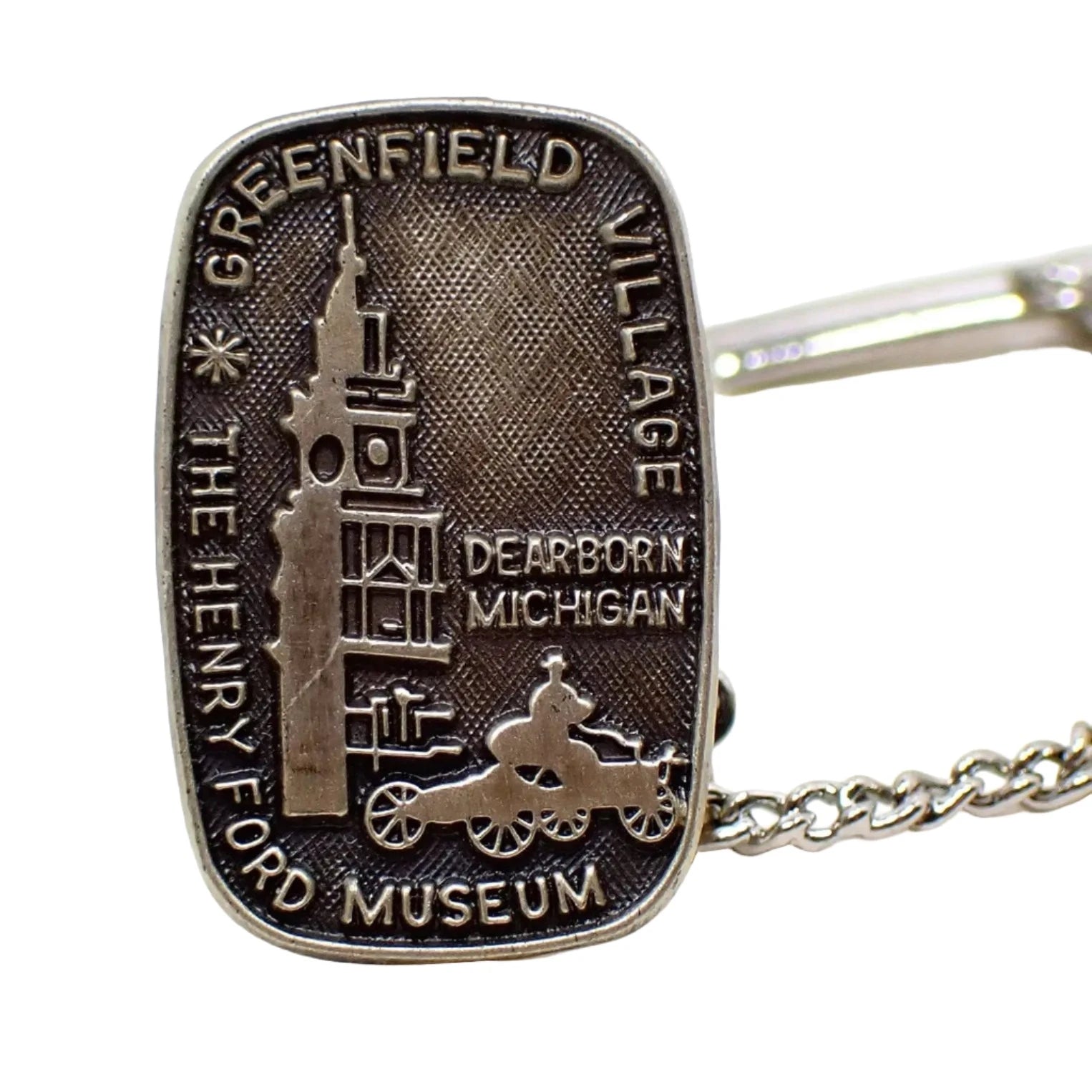 Enlarged front view of The Henry Ford Museum tie tack lapel pin. It has a rounded rectangle shape and has Greenfield Village and Dearborn Michigan on the front along with a depiction of the building and a man driving a Model T. The metal is antiqued sterling in color that has shades of gray. 