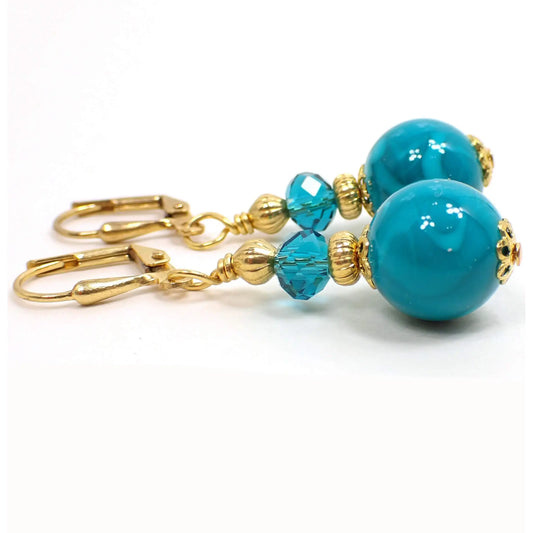 Side view of the handmade drop earrings. The metal is gold plated in color. There are teal blue faceted glass crystals at the top and marbled teal blue acrylic round beads at the bottom.