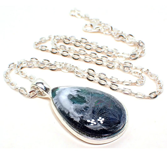 Enlarged angled front view of the handmade Frosty Water resin pendant. The metal is silver plated in color. The pendant is teardrop shaped and has iridescent blue at the bottom to an iridescent green on the top. There is an abstract frost like design in white through the resin.