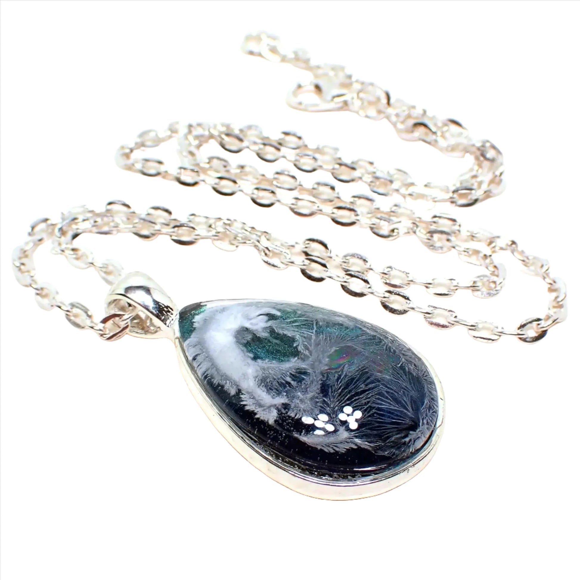 Enlarged angled front view of the handmade Frosty Water resin pendant. The metal is silver plated in color. The pendant is teardrop shaped and has iridescent blue at the bottom to an iridescent green on the top. There is an abstract frost like design in white through the resin.
