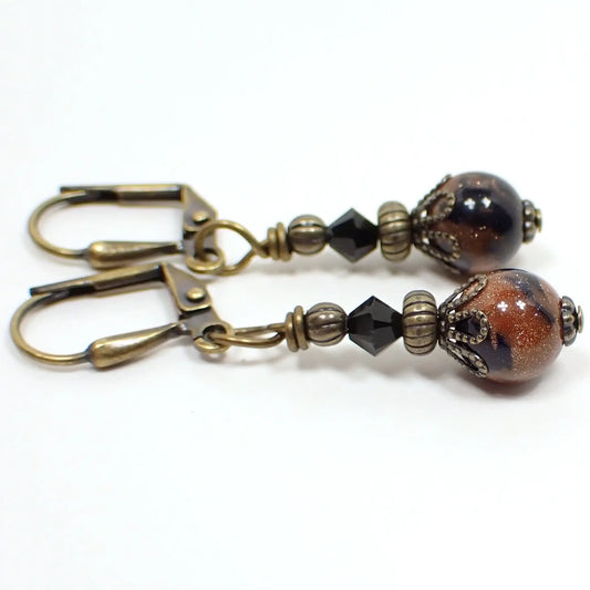 Side view of the handmade goldstone drop earrings. They have antiqued brass beads and findings. There are faceted black crystal glass bicone beads at the top and small round ball marbled goldstone and black beads at the bottom. The goldstone areas are orange with tiny flecks of copper.
