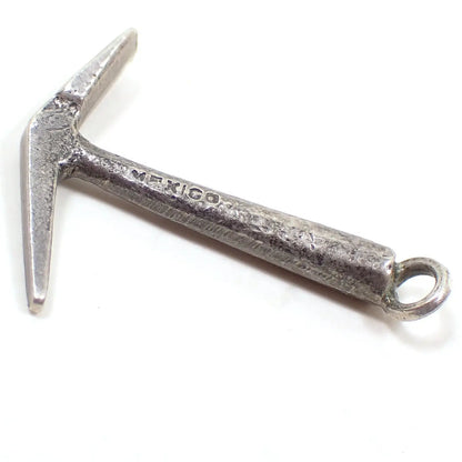 Enlarged side view of the small pick axe charm. It is silver tone in color and shaped like a small pick axe. It has Mexico stamped on the side of the handle part.