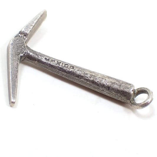 Enlarged side view of the small pick axe charm. It is silver tone in color and shaped like a small pick axe. It has Mexico stamped on the side of the handle part.