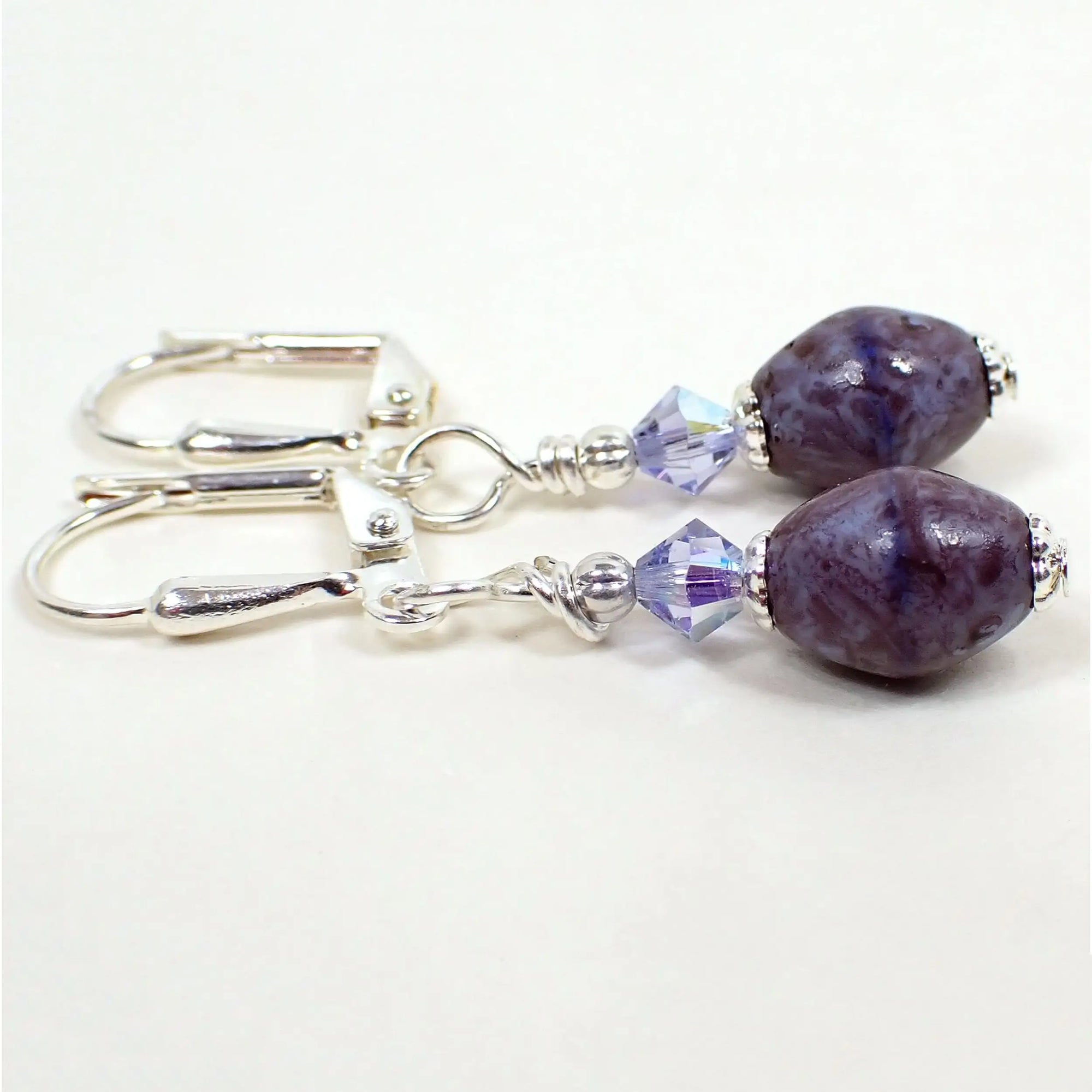 Side view of the small handmade drop earrings. The metal is silver plated in color. There are small faceted glass crystal beads at the top in a light purple color. The bottom beads are oval Czech glass beads and have mottled shades of purple and dark purple.
