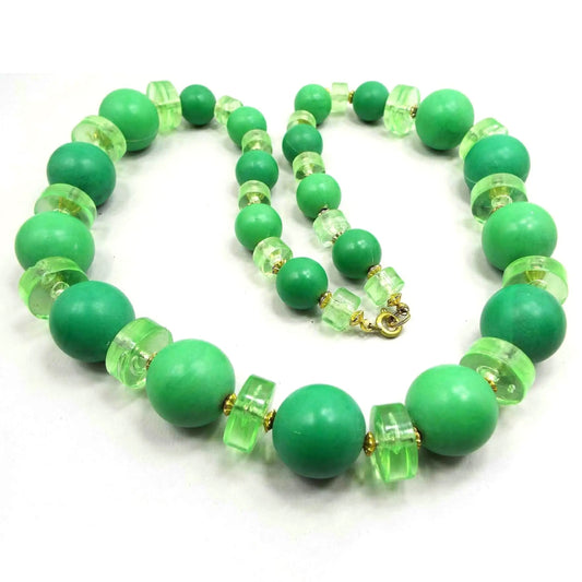 Top view of the Mid Century vintage chunky beaded necklace. It has large round green beads with translucent wide disc beads in between in varying shades of lime green. The metal beads and spring ring clasp are gold tone in color.