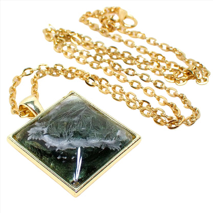 Enlarged front view of the handmade square abstract pendant necklace. The metal is gold plated in color. The resin is domed square shape with iridescent green that's most shades of olive green with hints of golden yellow. There is a white frost like design through the resin.