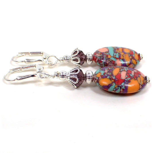 Side view of the handmade oval drop earrings. The metal is silver plated in color. The bottom beads are puffy oval shaped and have small blocks of different colors such as blue, pink, purple, red, and orange. There is a choice of top crystal glass bead in purple, orange, or yellow, but the ones in this photograph are purple in color.