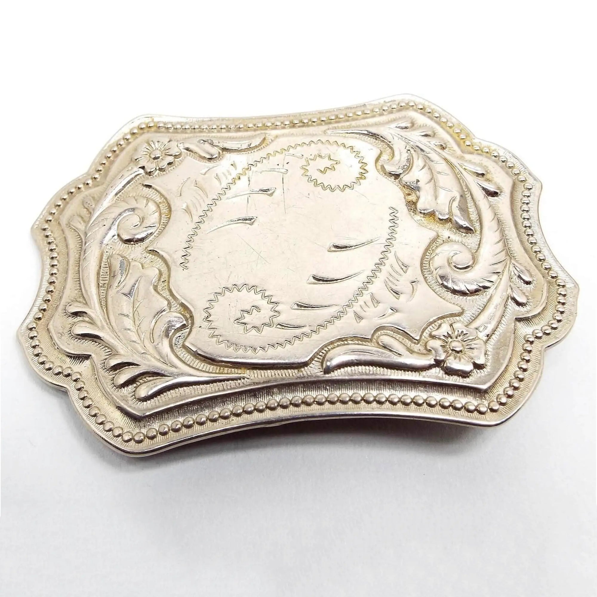 Front view of the retro vintage silver tone belt buckle. It has an etched and raised curled leaf like design and a dotted pattern around the edge. There is a flower on two of the corners. A few small scuff scratches from age can be seen on the front.