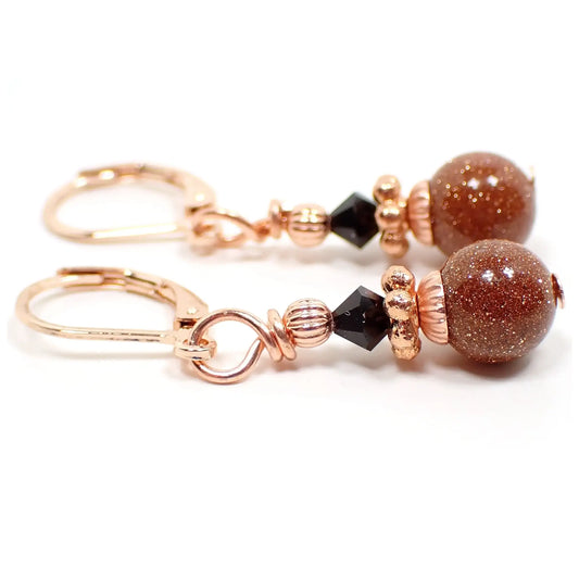 Side view of the handmade goldstone drop earrings. They have bright copper color and rose gold color beads and findings for a copper with hint of pink design. There are faceted black crystal glass bicone beads at the top and small round ball goldstone beads at the bottom. The goldstone beads are orange glass with tiny flecks of copper for sparkle.