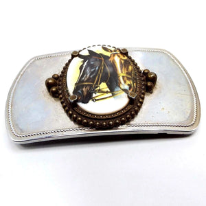 Front view of the retro vintage horses belt buckle. It is silver tone in color. The middle has a bezel frame that is antiqued brass in color. It is holding a white glass cab with a decal depicting two horse heads on it.