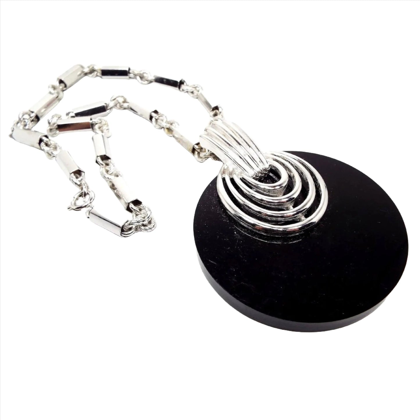 Angled view of the large black pendant necklace. The metal is silver tone in color. The chain has rectangle and textured round links. The pendant is a large black plastic circle that has an silver tone oval rings design at the top and a large silver tone color bail.