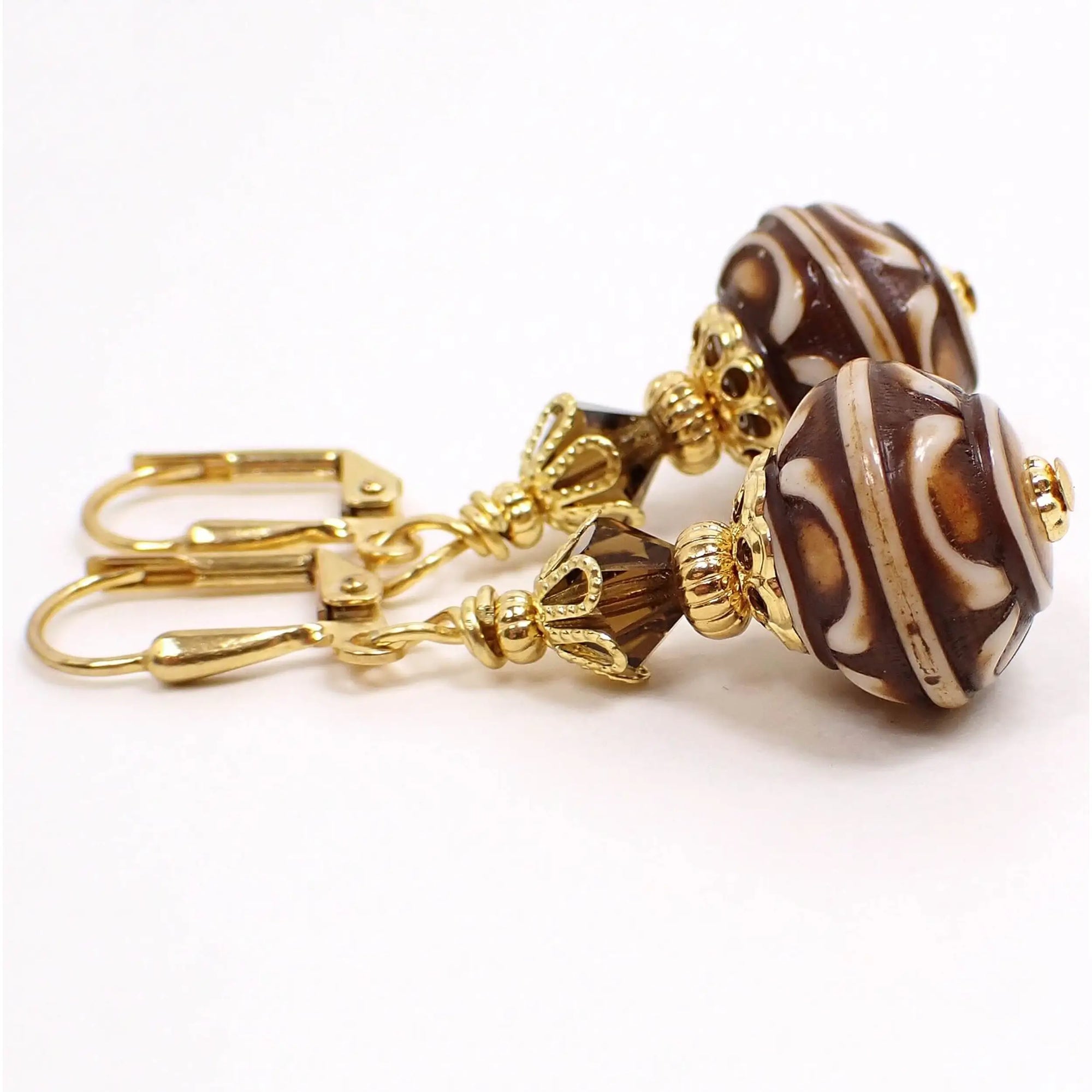 Side view of the handmade acrylic lantern earrings. The metal is gold tone in color. There are faceted brown glass crystal beads at the top. The bottom beads are acrylic and are saucer shaped with dark brown and cream color textured pattern.