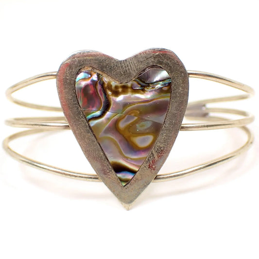 Enlarged front view of the retro vintage Mexican heart bracelet. The metal is silver tone in color. There are three rounded wires going from the back to the heart in the middle that form the cuff part of the bracelet. The heart has inlaid abalone shell. There are two pieces of shell. You can see the line in between the two pieces in the photo when viewed closely.