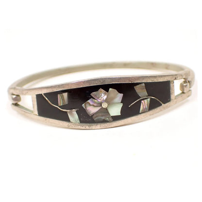 Front view of the Taxco retro vintage floral hinged bangle bracelet. The silver tone metal is slightly darkened from age. There is black enamel on the front with a flower design that has inlaid abalone shell for the flower petals and leaves.