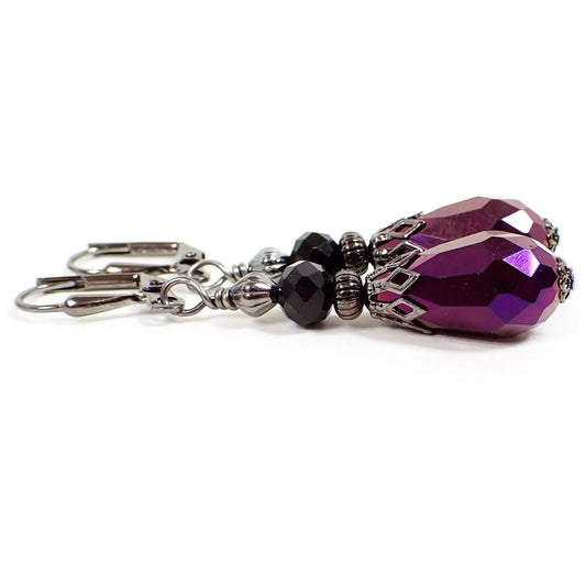 Angled view of the handmade teardrop earrings. The metal is gunmetal gray in color. There is a black faceted glass bead at the top. The bottom bead is a glass teardrop with a bright metallic purple color.