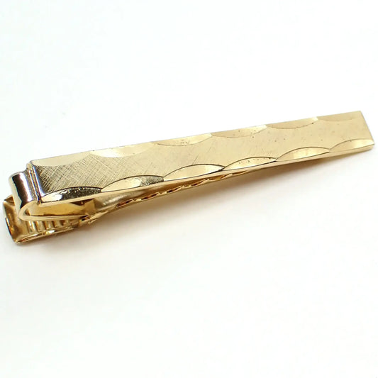 Angled top view of the retro vintage tie clip. The metal is brushed matte gold tone in color on the front. It is long rectangle shaped and has scalloped facets along the top and bottom edge.