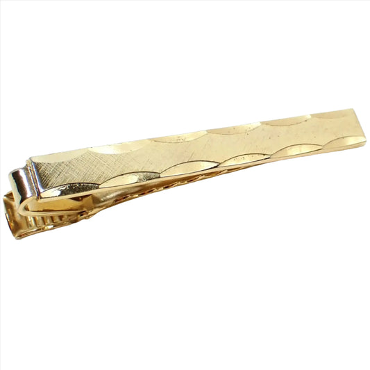 Angled top view of the retro vintage tie clip. The metal is brushed matte gold tone in color on the front. It is long rectangle shaped and has scalloped facets along the top and bottom edge.
