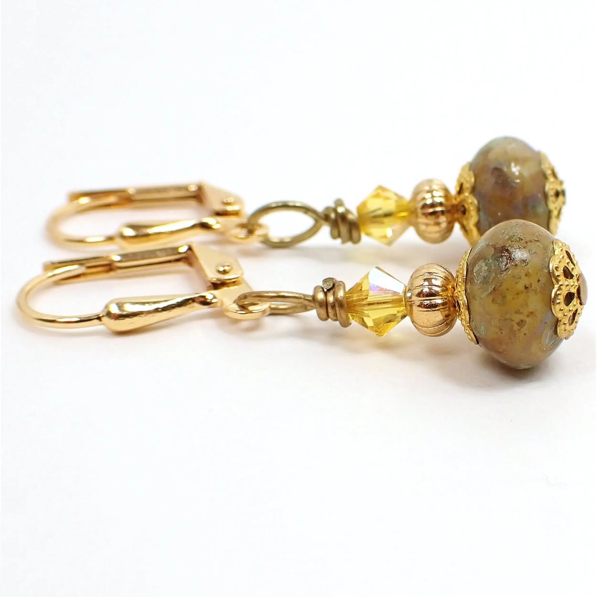 Side view of the handmade beaded glass drop earrings. The metal is gold plated in color. There is a yellow faceted glass crystal bead at the top of the earrings. The bottom glass Czech beads have mottled mustard yellow tones with some specks of green for an overall dark yellow appearance.