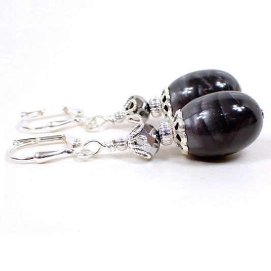 Angled view of the handmade drop earrings with vintage lucite beads. The metal is silver plated in color. There are faceted metallic silver glass beads at the top. The bottom lucite beads are oval shaped and have a pearly mix of light and dark gray color.