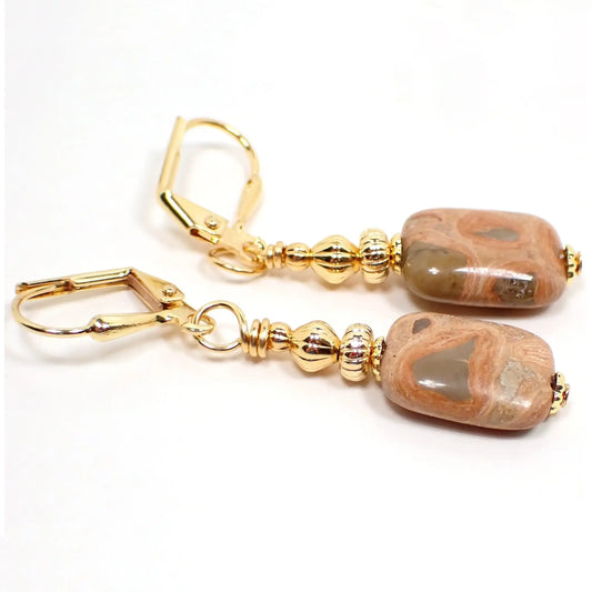 Angled view of the handmade safari jasper earrings. There are lever back earwires at the top in this photo. The metal is gold plated in color. The gemstones are puffy oval shaped and have marbled areas of soft green, brown, peach, and orange.