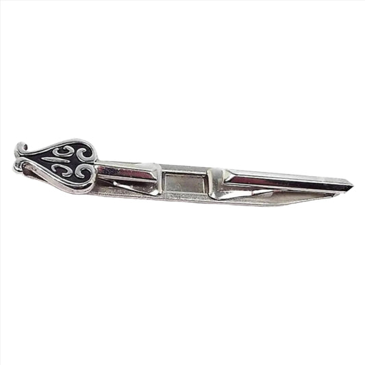 Front view of the Mid Century vintage Swank tie bar. The metal is silver tone in color. There is an open area in the middle for the ends to slide over each side of the tie. One end has a spade like design with black paint.