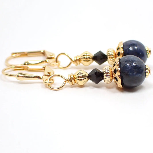 Side view of the small handmade sodalite gemstone earrings. The metal is gold plated in color. There are black faceted glass crystal beads at the top. The bottom gemstone beads are round sphere shaped and mostly dark denim blue in color with specks of black and gray.
