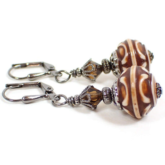 Side view of the handmade lantern drop earrings with acrylic beads. The metal is gunmetal gray in color. There are brown faceted glass crystal beads at the top. The bottom beads are acrylic and are round saucer shaped with a cream and brown raised design.